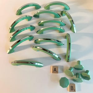 May include: A collection of twelve arched mint green cabinet pulls with metallic silver trim, accompanied by four matching octagonal mint green cabinet knobs. Two small wooden tiles labelled A1 and B3 are positioned near the cabinet hardware.