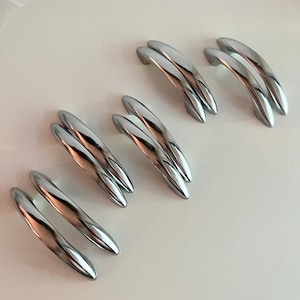 Peut inclure: Un ensemble de dix poignées de placard en métal argenté disposées par paires. Chaque poignée présente une forme arquée et lisse en croissant avec une finition polie et réfléchissante. Les poignées sont placées en diagonale de manière uniforme.