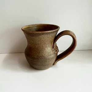 May include: A handmade brown ceramic mug with a curved handle. The mug has a textured surface and a wide opening. The colour is a mix of brown and tan, giving it a rustic appearance. The mug is suitable for hot or cold beverages.