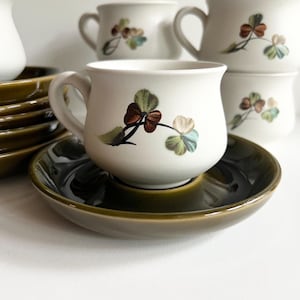 May include: A set of white ceramic teacups with green and brown shamrock designs. The cups are paired with matching green saucers.