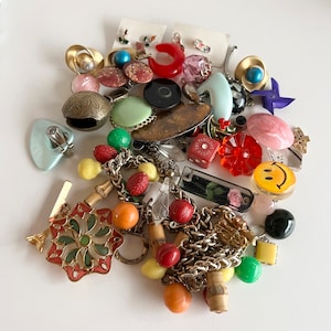 Puede incluir: Una colecci&oacute;n de abalorios y cuentas vintage de colores, que incluyen un abalorio de cara sonriente, un abalorio de coraz&oacute;n rojo, una cuenta a rayas verde y blanca y una cadena dorada. Los abalorios est&aacute;n dispuestos sobre una superficie blanca.