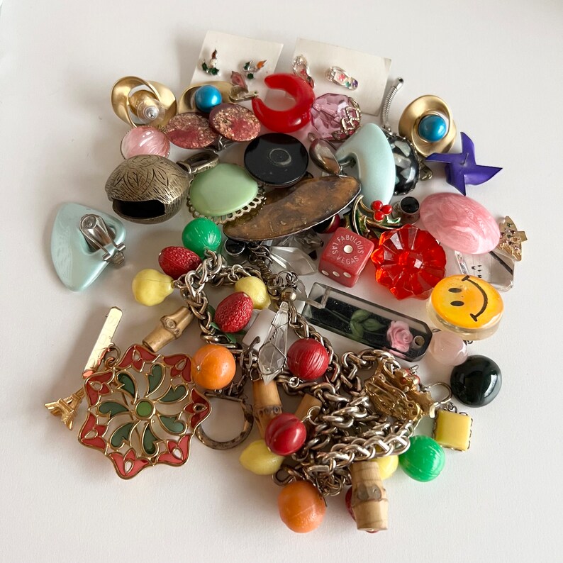 Puede incluir: Una colecci&oacute;n de abalorios y cuentas vintage de colores, que incluyen un abalorio de cara sonriente, un abalorio de coraz&oacute;n rojo, una cuenta a rayas verde y blanca y una cadena dorada. Los abalorios est&aacute;n dispuestos sobre una superficie blanca.