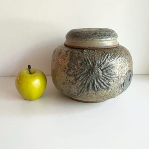 May include: A ceramic jar with a lid, featuring a textured surface and a floral design. The jar is a mix of grey and brown tones. A green apple is next to the jar.