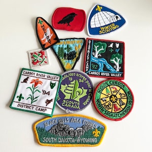 Könnte beinhalten: Eine Sammlung von Vintage-Scout-Abzeichen, darunter ein schwarzer Rabe auf einem roten Abzeichen, ein weißer Iglu mit dem Text "Winter Scouting", ein grün-weißes Abzeichen mit einer Lilie und dem Text "Carrot River Valley District Camp", ein violettes Abzeichen mit dem Text "Scouts Canada" und ein gelbes Abzeichen mit dem Text "Black Hills Area Council South Dakota Wyoming".