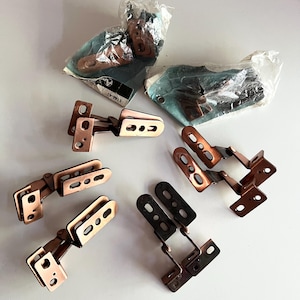 May include: A collection of copper-coloured metal hinges with multiple elongated mounting slots. Some hinges are loose, while others remain inside clear plastic packaging. The packaging includes a small label with the text 5146-41 visible.