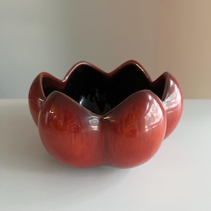May include: A glossy, deep red ceramic bowl with a scalloped, flower-like rim. The exterior features a rich, variegated red glaze with darker undertones, while the interior is finished in a smooth, solid dark brown or black glaze.