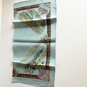 May include: A light blue silk scarf with a red, green and yellow tartan border. The scarf features a watercolour illustration of a ship sailing past a mountain range with the text "The Waters of Skye" and "Castle Campbell and the Falls of Dochart" printed on the scarf.