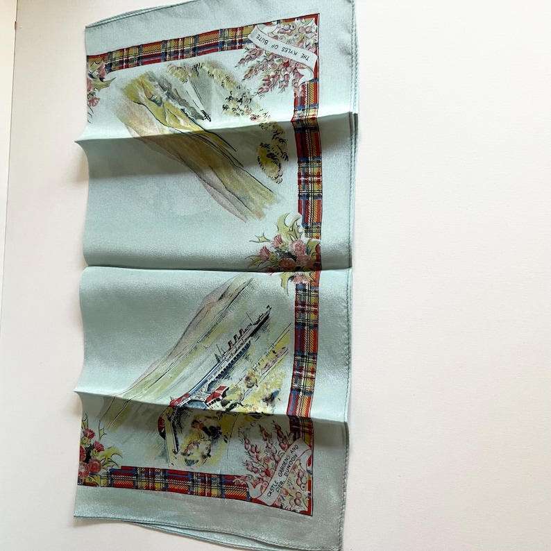 May include: A light blue silk scarf with a red, green and yellow tartan border. The scarf features a watercolour illustration of a ship sailing past a mountain range with the text "The Waters of Skye" and "Castle Campbell and the Falls of Dochart" printed on the scarf.
