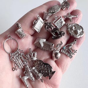 May include: A collection of silver charm pendants, including a house, an owl, a crown, a suitcase, a palm tree, a radio, a shell, a horseshoe, a plane, a woman with a bucket, a key, and a mug.