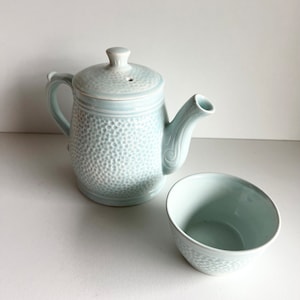 Könnte beinhalten: Eine hellblaue Teekanne und eine passende Teetasse aus Keramik mit einem strukturierten, gesprenkelten Design.