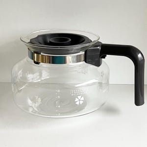 May include: A clear glass coffee pot with a black plastic lid and handle. The pot has a silver metal band around the top and markings for 2, 4, 6, and 8 cups. The text "TENS & CUPS TO BOTTOM OF METAL BAND" is printed on the side of the pot.