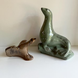 May include: Two ceramic seal figurines. One is a seated, glossy, sage green seal with a raised head. The other is a smaller, brown seal figurine in a reclining position. Both are on a white surface.