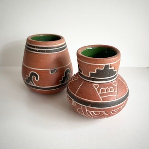 May include: Two small, brown ceramic pots with black and white geometric designs. The pots have a green glaze on the inside.