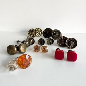 May include: Assortment of vintage cabinet knobs in various colors and materials, including brass, glass, and plastic. The knobs feature a variety of designs, including floral, geometric, and abstract patterns.