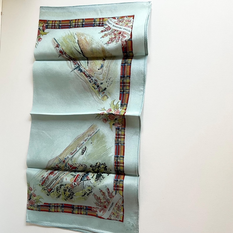 May include: A light blue silk scarf with a plaid border and a floral design. The scarf features a watercolour-style illustration of a Scottish landscape with a castle, a river, and a bridge. The text "SCOTTISH HIGHLANDS" is printed on the scarf.