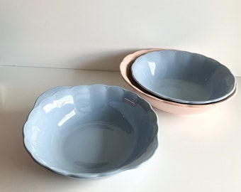 May include: Two blue ceramic bowls with scalloped edges, one nested inside the other, on a white surface. The larger bowl is sitting on top of a smaller pink bowl.