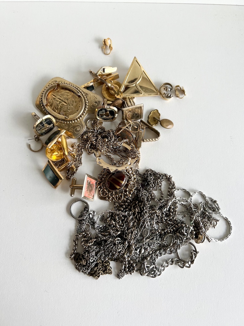Puede incluir: Una colecci&oacute;n de joyas de oro y plata, que incluye cadenas, pendientes, broches y gemelos. Las joyas est&aacute;n dispuestas en una pila sobre una superficie blanca.
