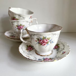Könnte beinhalten: Ein Set aus weißen Porzellantassen und Untertassen. Jede Tasse und Untertasse hat ein Blumendesign mit rosa Rosen und blauen Akzenten. Die Ränder sind mit goldenen und grauen Details verziert. Die Tassen sind gestapelt, wobei eine Tasse und Untertasse im Fokus stehen.