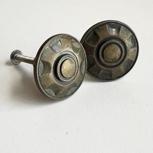 Peut inclure: Deux boutons de meuble anciens en laiton avec un design circulaire. Chaque bouton présente un cercle central entouré d'anneaux concentriques et d'un bord extérieur décoratif. Un bouton a une vis attachée pour l'installation. Les boutons ont un aspect vintage et vieilli.