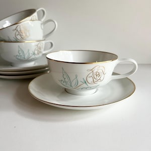May include: A set of four white teacups and saucers with a gold rim. The cups and saucers feature a delicate floral design with a gold outline and green leaves.
