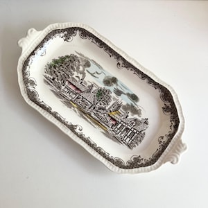 May include: A vintage rectangular serving tray with a detailed landscape scene in the center. The tray has a white base with a black and brown patterned border and decorative handles. The landscape features buildings, trees, and a church.