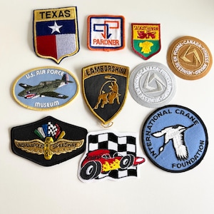 May include: A collection of ten embroidered patches featuring various designs, including a state flag, a car logo, a military emblem, and a sports team logo. The patches are all different colors and sizes, and they are all sewn onto a white background.