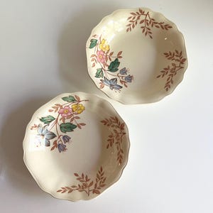 May include: Two white ceramic bowls with scalloped edges and floral designs. The bowls feature a brown leafy pattern and colourful flowers in shades of pink, yellow, blue, and green.