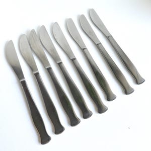 May include: Eight silver butter knives with rounded handles. The knives are arranged in a row, with the handles pointing to the right.