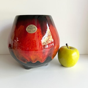 May include: A glossy, red, black, and orange ceramic vase with a rounded shape and a small base. The vase has a label that reads "Sofia Originals Limited Toronto Canada". A green apple sits next to the vase.