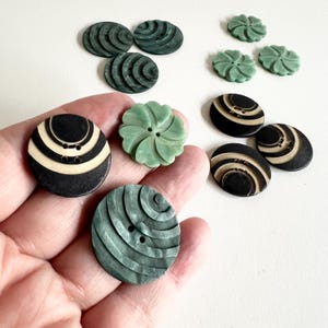 May include: A collection of vintage buttons in various designs and colors. The buttons include dark green, black, and cream-colored options. Some buttons have a spiral design, while others are flower-shaped or feature striped patterns. The buttons are displayed on a white surface.