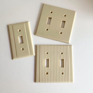 May include: Three vintage light switch plates in a cream colour. Two of the plates have a smooth surface and two toggle switches. The third plate has a textured surface and two toggle switches.