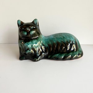 May include: A ceramic cat figurine in a reclining pose. The cat has a glossy finish with a blend of teal, black, and brown colors. The figurine is a decorative item, likely for display.