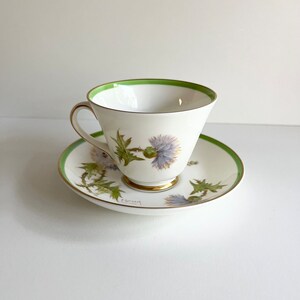 May include: A white teacup and saucer with a green rim and a floral design featuring purple thistles. The saucer has a gold rim and the teacup has a gold band around the base. The teacup and saucer are signed "P. Gormack".