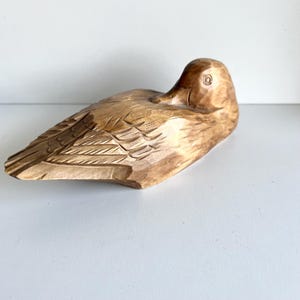 May include: A wooden sculpture of a duck in a resting pose. The duck is carved from light brown wood, with detailed feather and eye carvings. The sculpture is on a white surface.