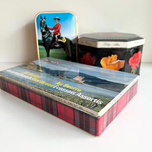 May include: Three vintage metal tins. One rectangular tin with a landscape scene and red plaid sides. A second tin is a black octagon with floral designs. The third tin is a blue rectangle with a rider on a horse.