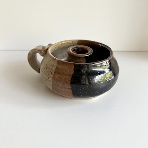 May include: A handmade ceramic candle holder with a handle. The candle holder is round with a central opening for a candle. The exterior features a color block design in shades of beige, brown, and black.