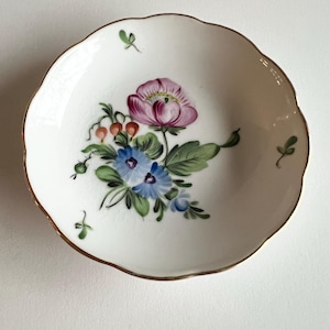 May include: A small, white, scalloped-edge dish with a hand-painted floral design. The flowers are a mix of pink, blue, and green, with a gold rim. The dish is likely porcelain or ceramic.