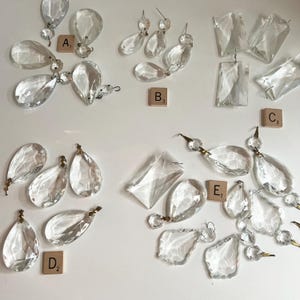 May include: A collection of clear glass chandelier crystals in various shapes, including teardrops and rectangles, organized into groups labeled A1, B3, C3, D2, and E1 with small wooden tiles. Each crystal features metal loops for hanging.