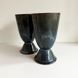 May include: Two handmade ceramic goblets. The goblets are dark blue with a glossy finish and a flared base. The interior of the goblets has a lighter blue and white color. The goblets are displayed on a white surface.