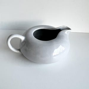 May include: White ceramic creamer or gravy boat with a rounded body and a curved spout. The handle is attached to the side of the body.
