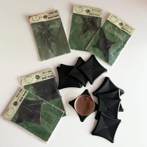May include: A collection of black Amerock back plates, some sealed in original green and white packaging labelled E588-18A, E513-18A, and E511-18A. Several loose, square-shaped black back plates with concave sides are arranged alongside a copper coin.