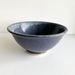 Peut inclure: Un bol en céramique fait à la main, de couleur indigo foncé avec une texture mouchetée. Le bord présente un glaçage gris-bleu contrastant, et la base est de couleur beige clair. Ce bol est idéal pour servir des aliments.