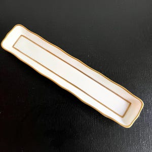 Peut inclure: Un plateau rectangulaire en céramique blanche avec une bordure dorée et un liseré doré. Le plateau a un bord légèrement ondulé et est vide. Le plateau est sur une surface noire.