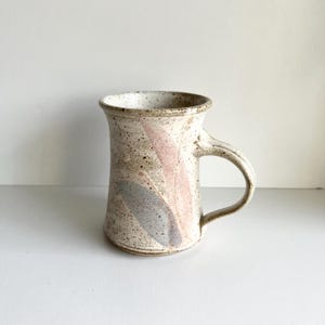 May include: A white ceramic mug with a speckled glaze and a pink and blue abstract design. The mug has a rounded body and a handle.