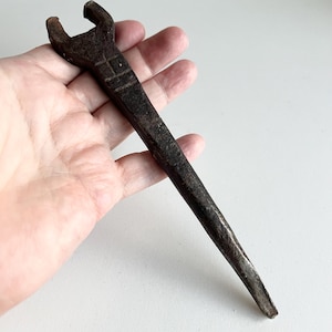 Peut inclure: Une clé en métal brun foncé d'époque, avec une tête en forme de fourche et un long manche effilé. La clé a une surface légèrement texturée et semble usée, suggérant l'âge et l'utilisation. Un petit marquage en forme de carré est visible sur le manche.