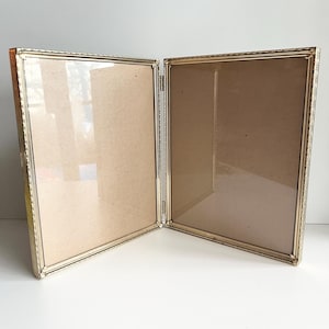 May include: A vintage, hinged double picture frame with a gold-colored metal frame. The frame is open, revealing two sections with glass and a tan backing. The frame is on a white surface.