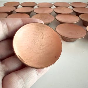 Könnte beinhalten: Eine Sammlung kleiner, kreisförmiger, kupferfarbener Metallscheiben mit einer leicht konkaven Oberfläche. Jede Scheibe weist eine glatte, metallische Oberfläche mit subtilen Farbnuancen auf und ist in einem Rastermuster auf einer flachen Ebene angeordnet.