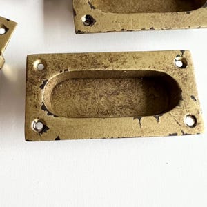 Peut inclure: Une poignée de tiroir rectangulaire dorée avec une découpe ovale. La poignée a une finition vieillie et deux petits trous de chaque côté.