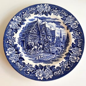 May include: A blue and white plate with a detailed illustration of a horse-drawn carriage in a European village. The plate has a floral border and a classic, vintage aesthetic.