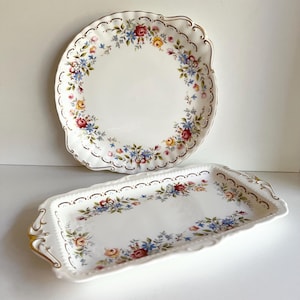 May include: Two white porcelain serving dishes with floral patterns. The round dish and rectangular tray feature a scalloped edge with gold trim and a design of red, blue, and yellow flowers.